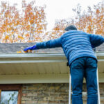 Man in ladder removing autumn leaves from gutter during day of autumn