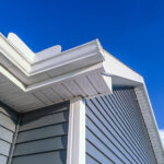 A low angle view of soffit, gutters, downspout and vinyl siding on a new home. Blue sky is in the background.