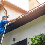 man on ladder cleaning house gutter from leaves and dirt